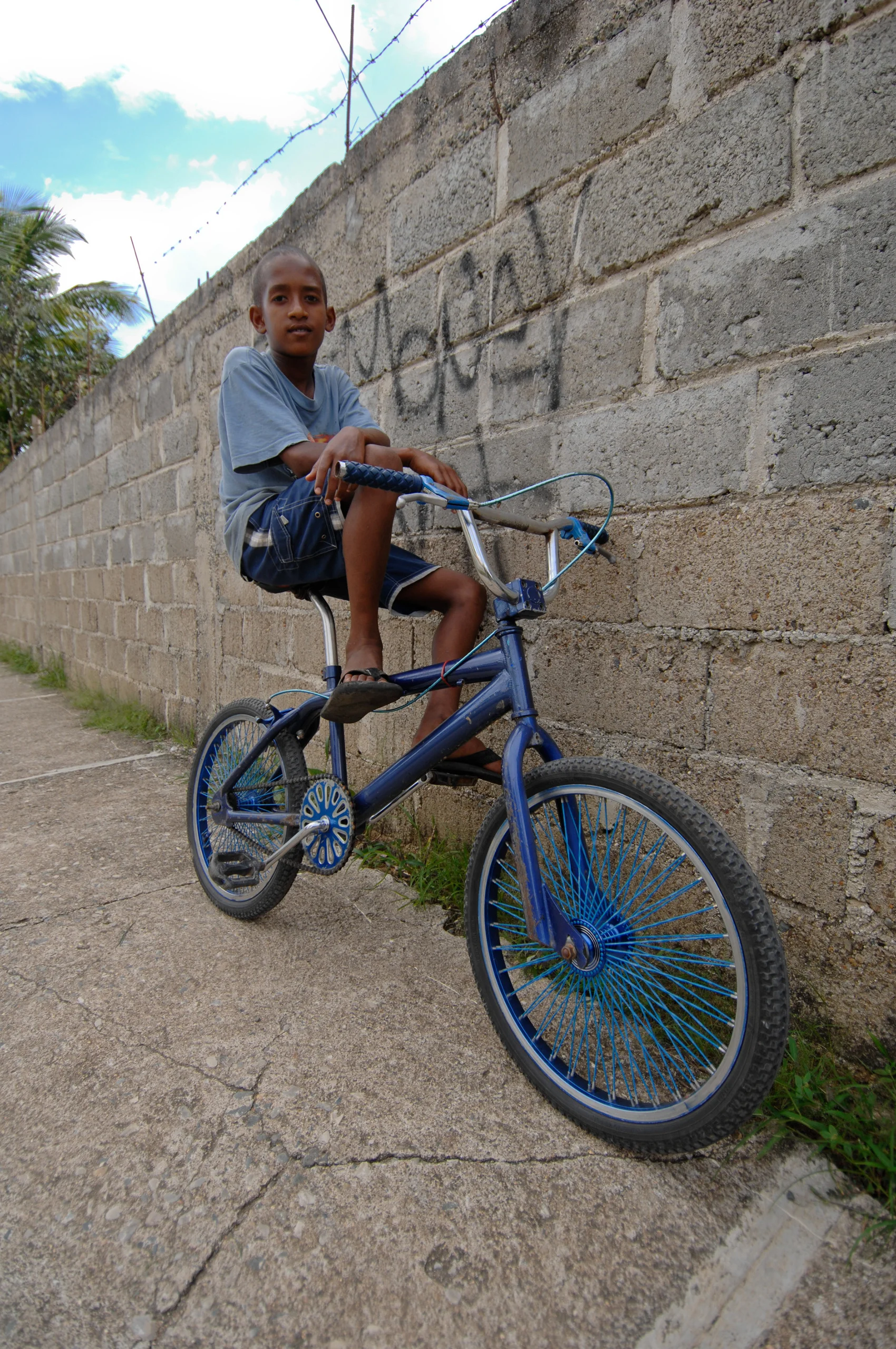 A person sitting on a blue bicycle beside a concrete block wall.