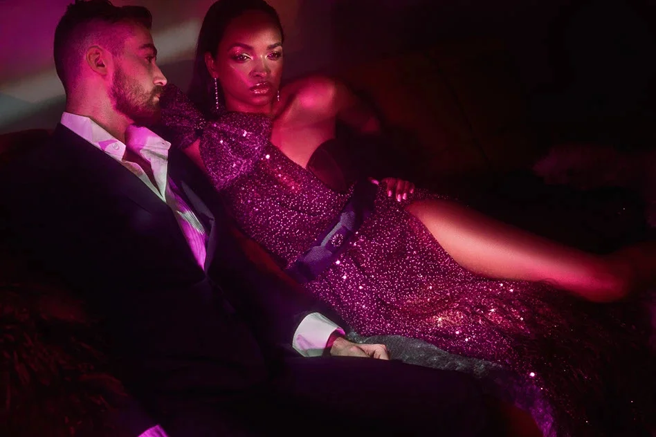 Two models reclining together on a sofa under red and magenta light.