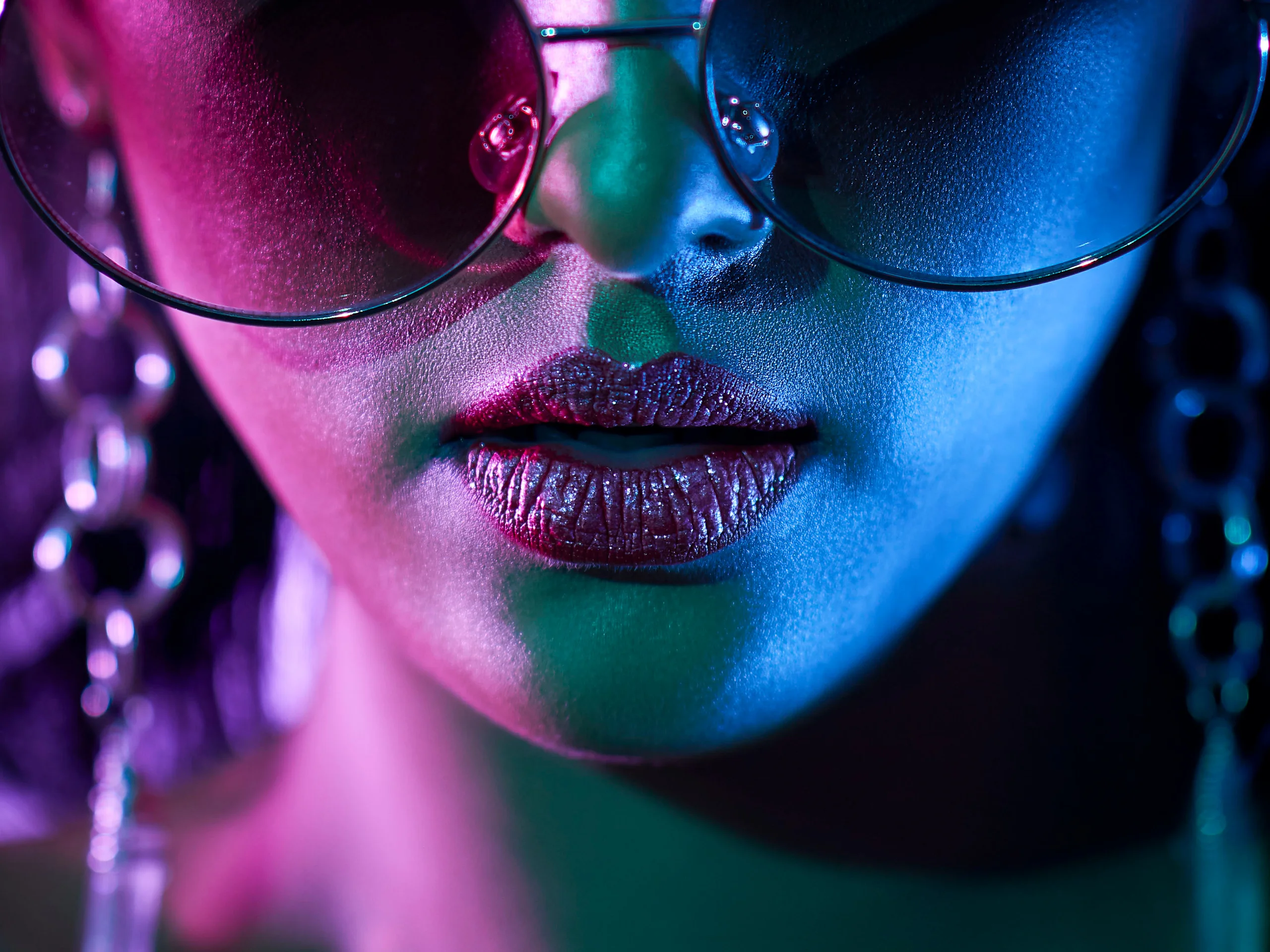 A close-up of oversized sunglasses and glossy lips under pink, green and blue light.
