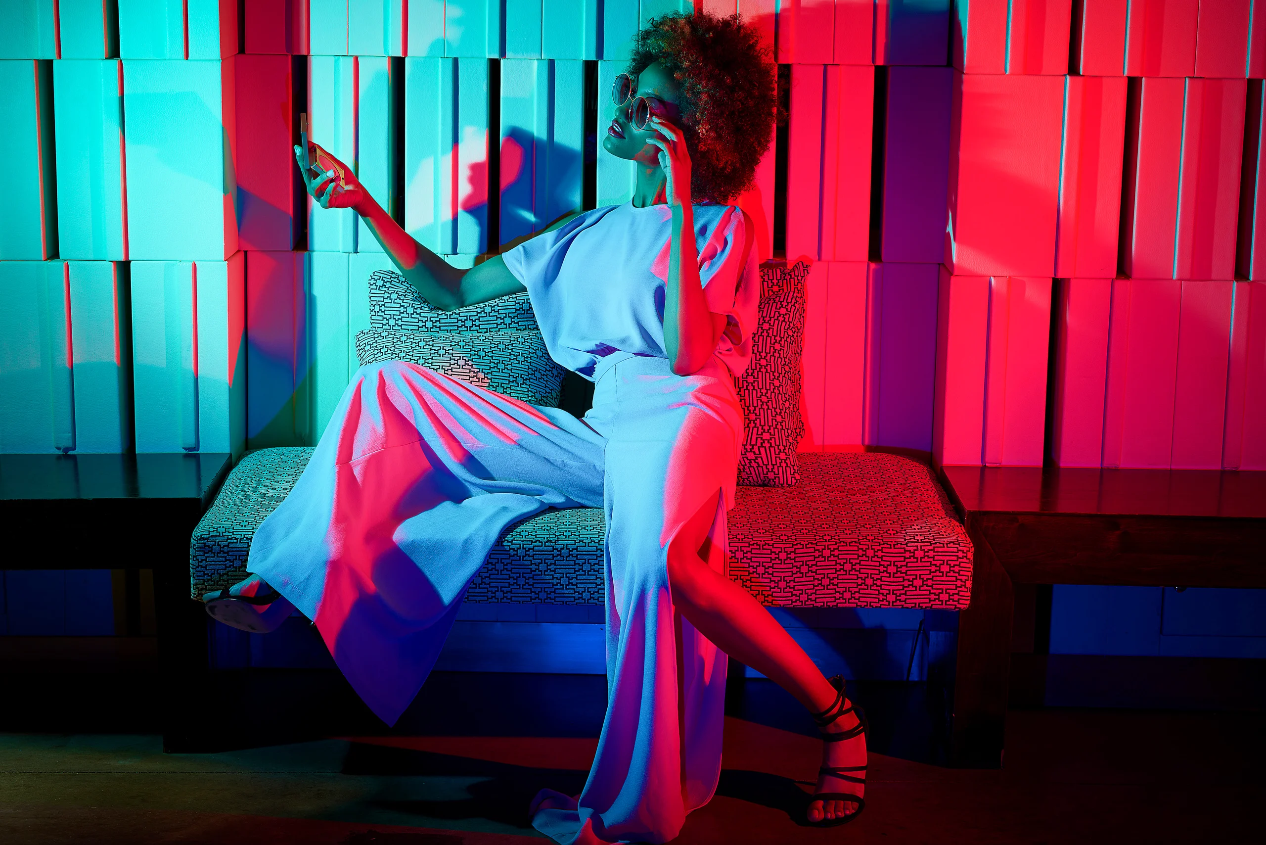 A model in a pale suit seated against a geometric wall under cyan and red light.