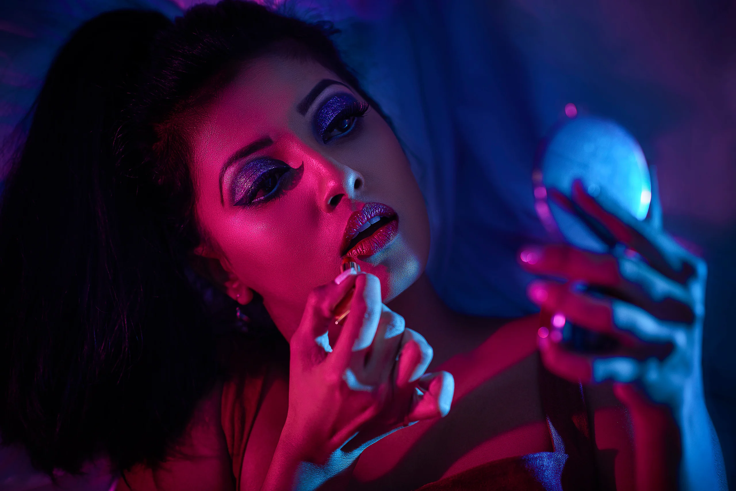 A model applying lipstick while looking into a handheld mirror under pink and blue light.