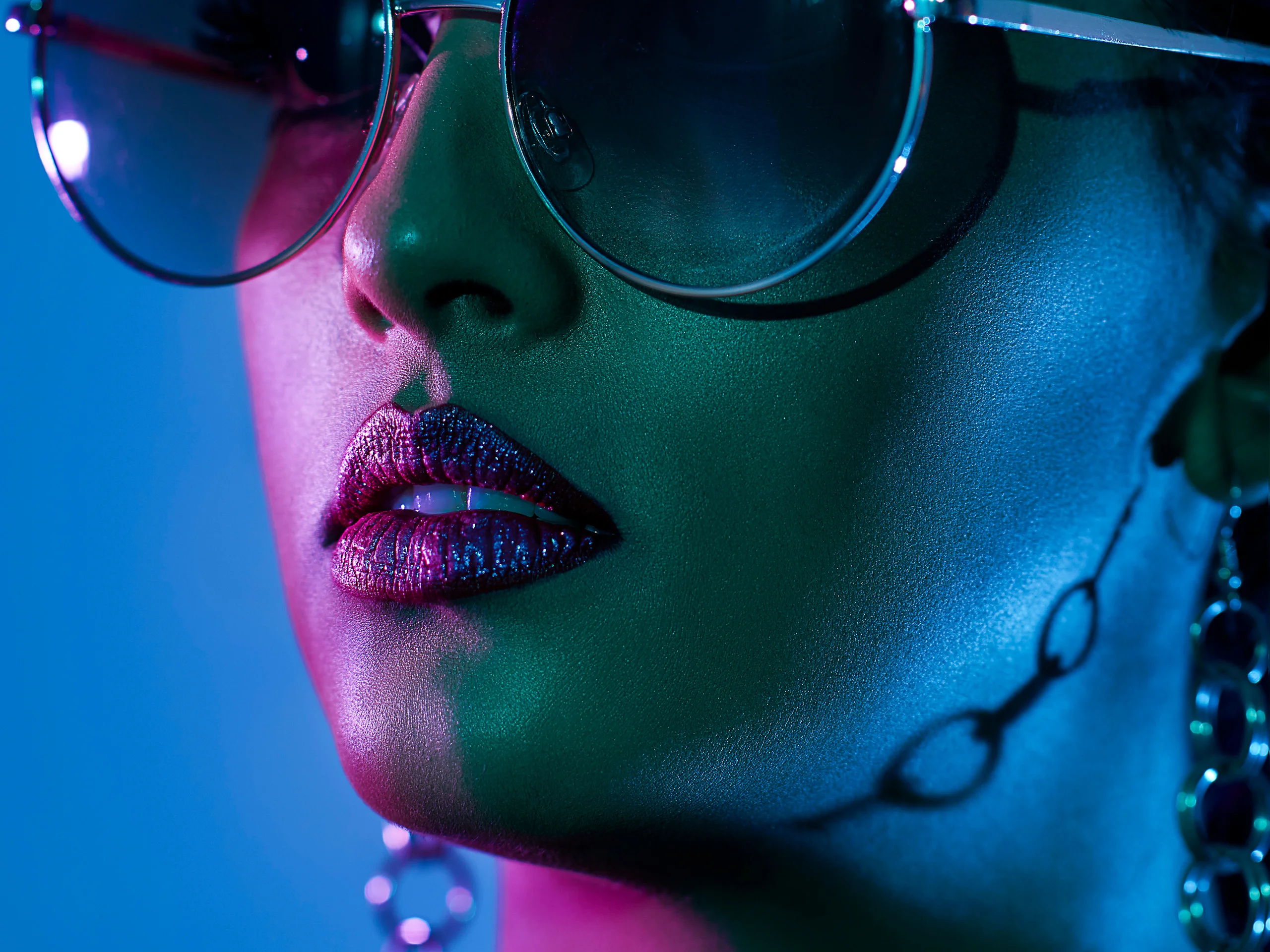 A close beauty portrait in oversized sunglasses with a chain across the cheek under blue and pink light.