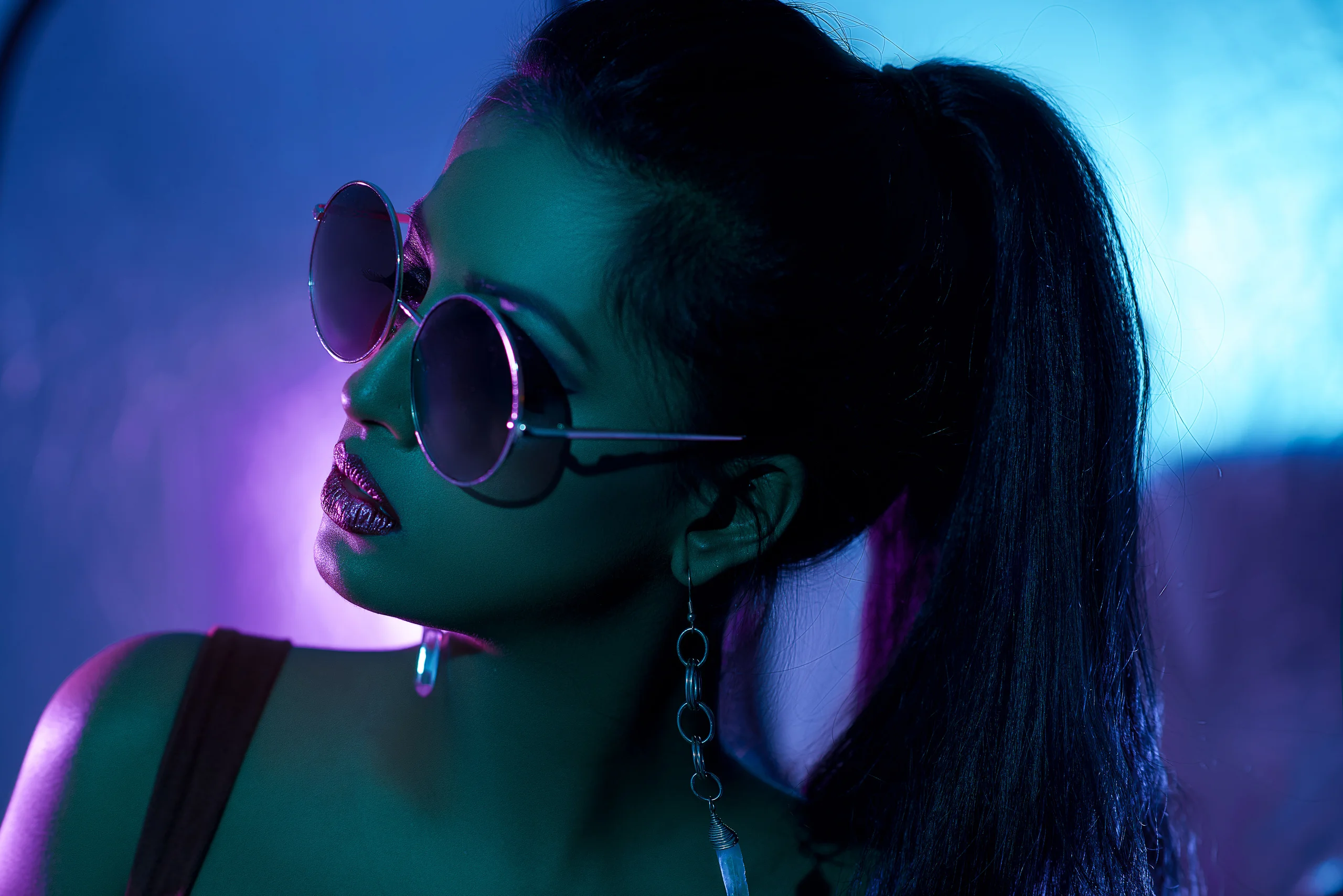 A model with a high ponytail and round sunglasses turned to the side under blue and purple light.