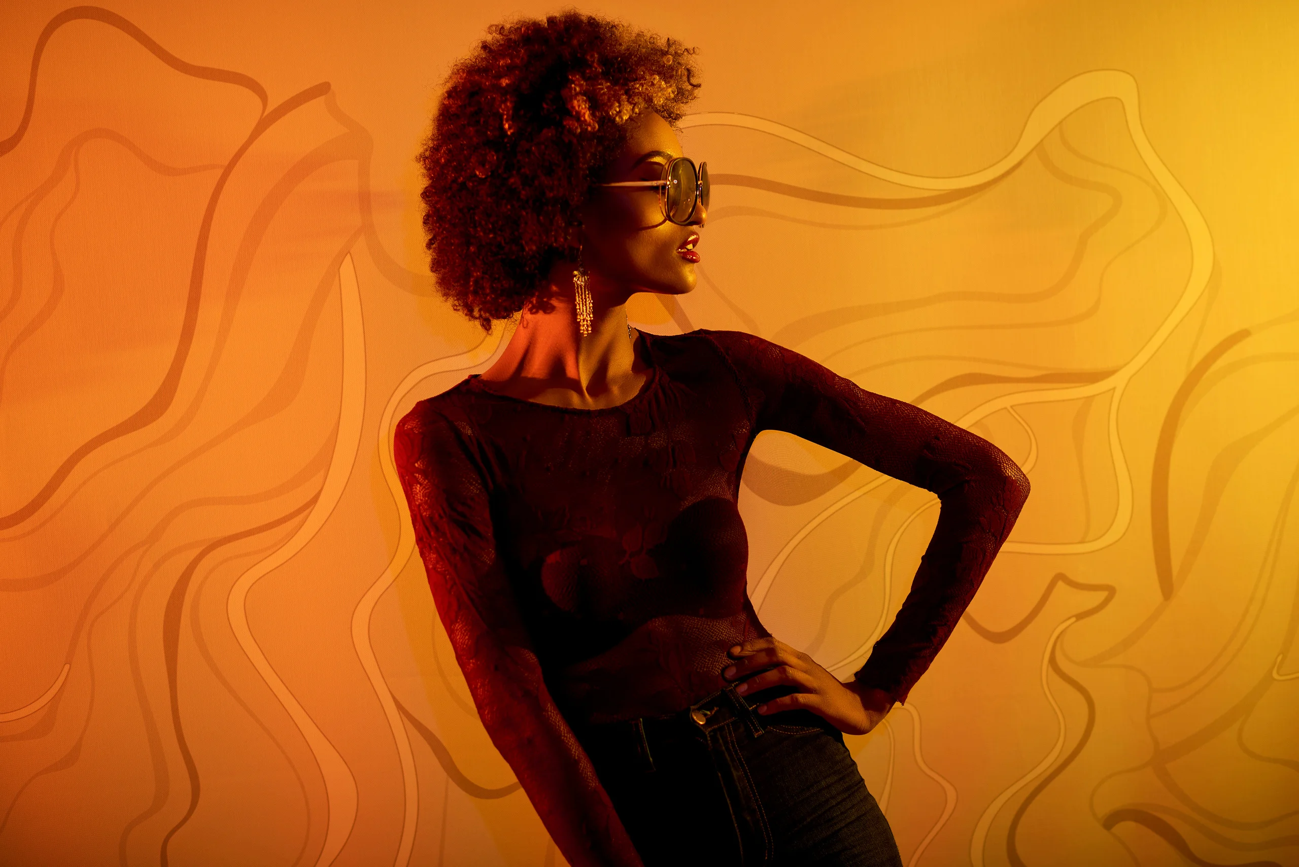 A model in sunglasses and a dark fitted outfit posing against an orange patterned wall.