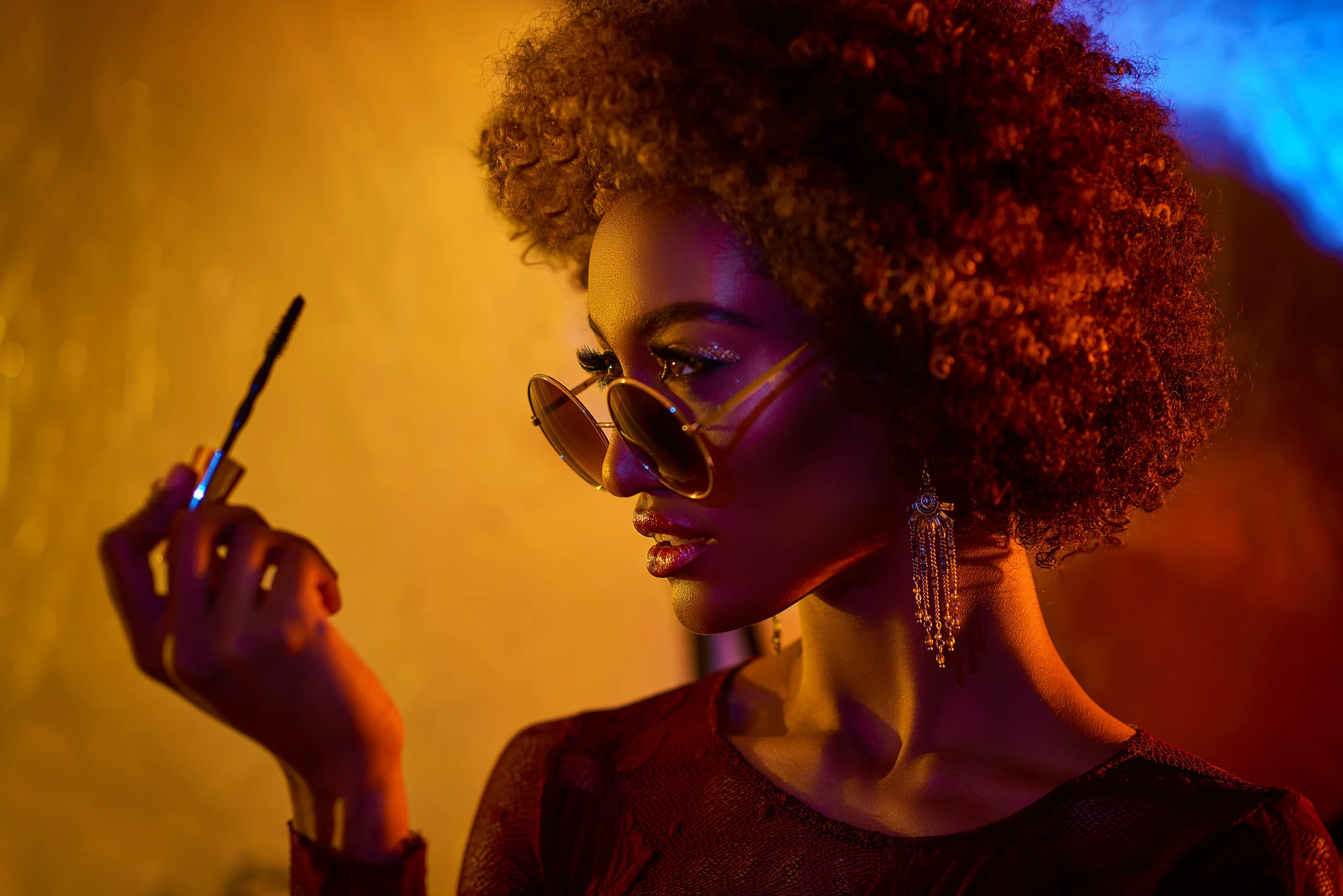 A model with curly hair and sunglasses examining a makeup brush under warm light.