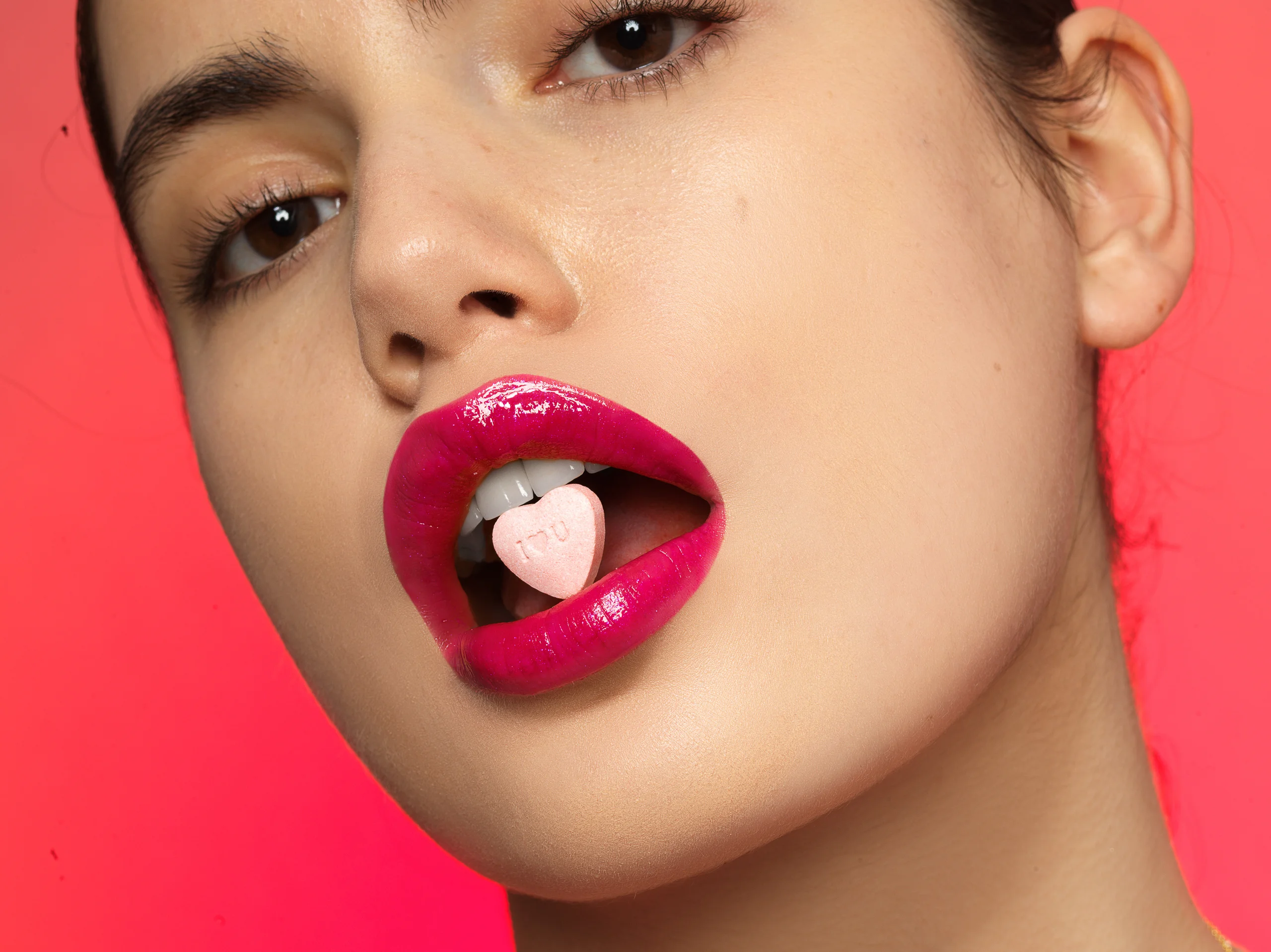 A close beauty portrait with vivid pink lip gloss and a small round sweet on the tongue.
