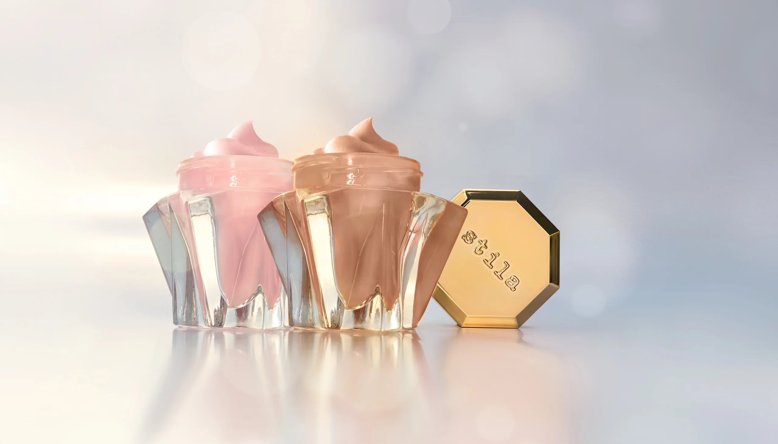 Stila Lingerie Soufflé cosmetic jars and their gold lids on a reflective surface.