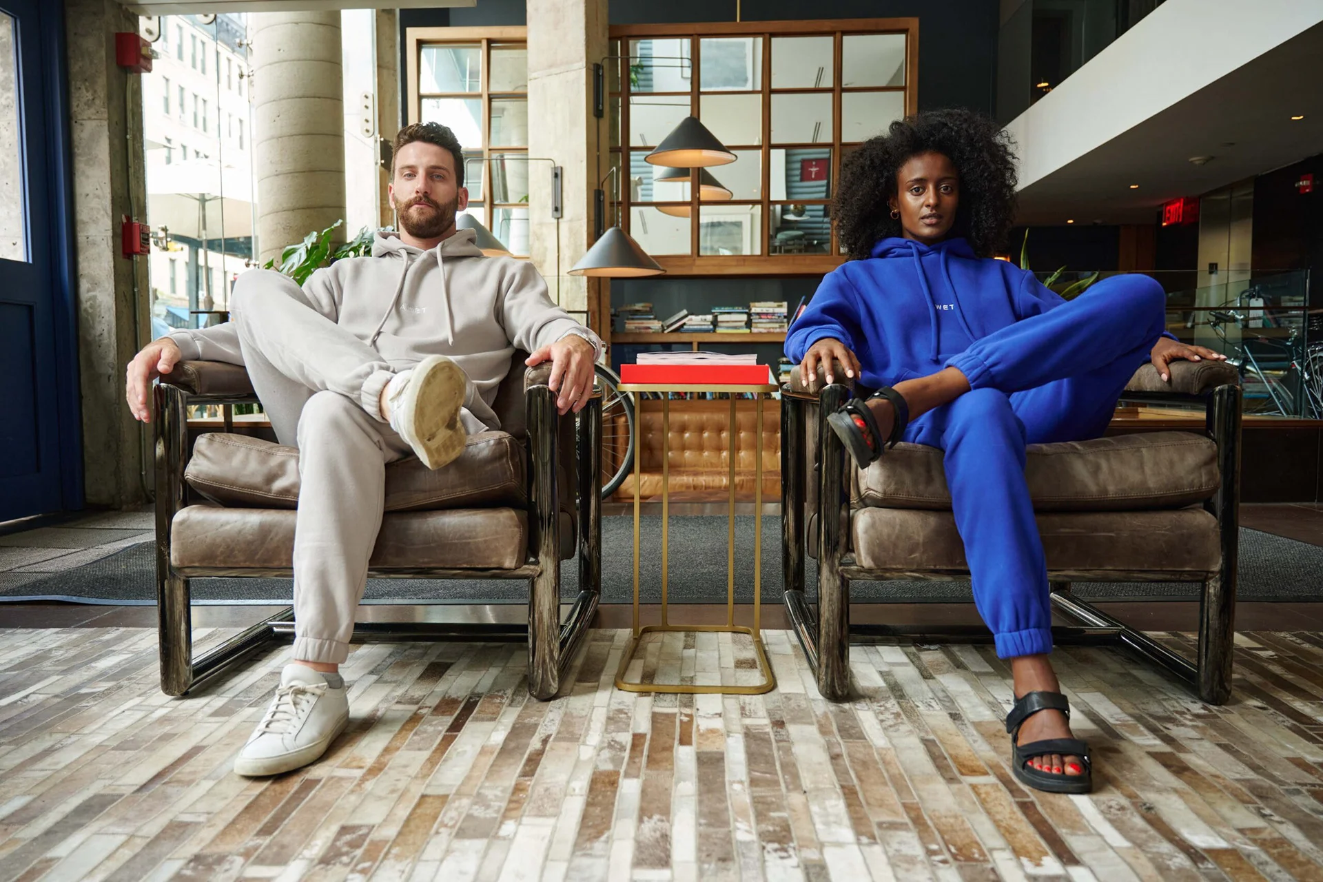 Two models in gray and blue ÁWET outfits sitting in leather armchairs.